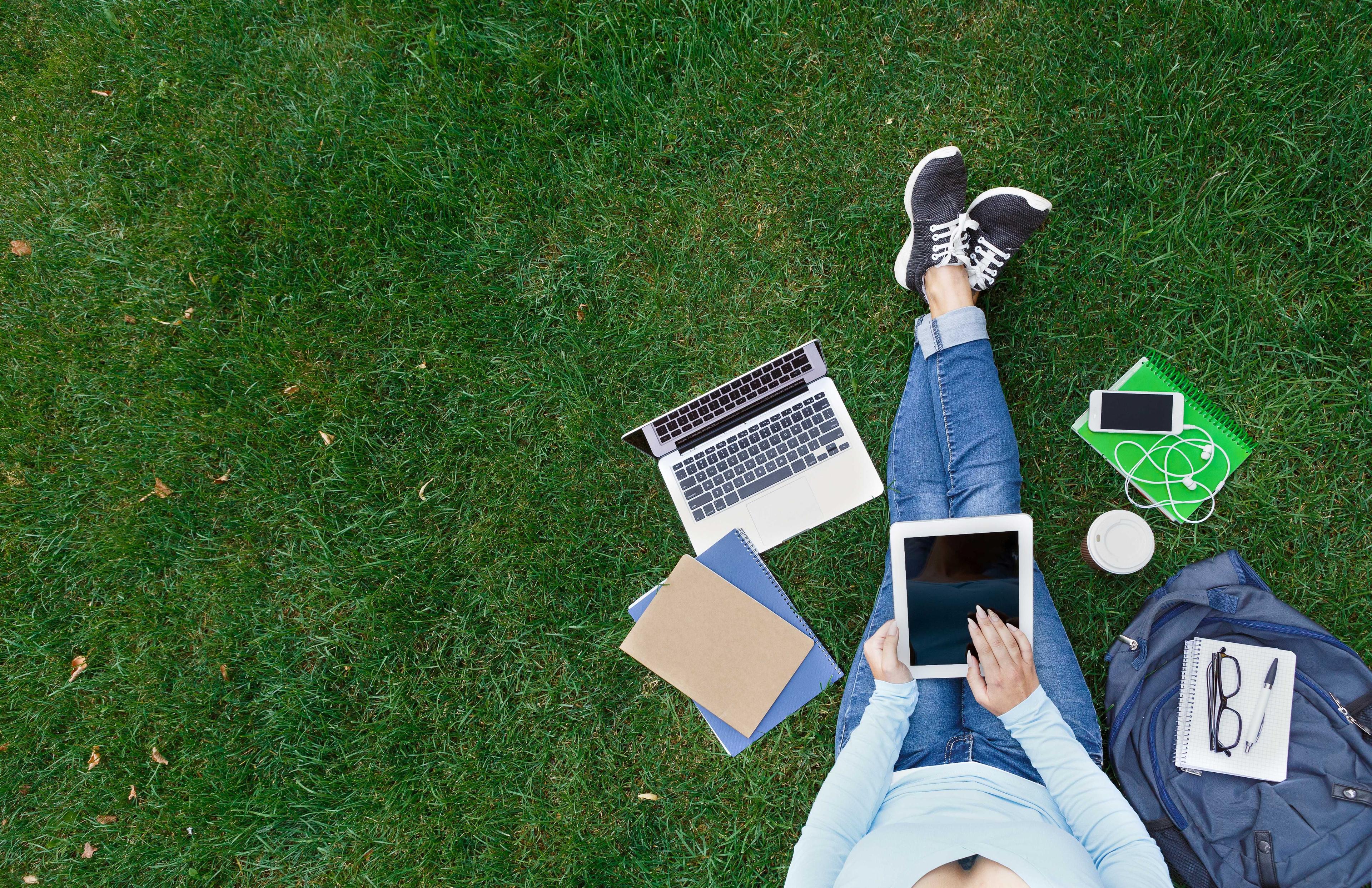 A woman sitting on the grass and holding a tablet. She is surrounded by different objects such as a laptop, a mobilephone, notebooks and a bag.
